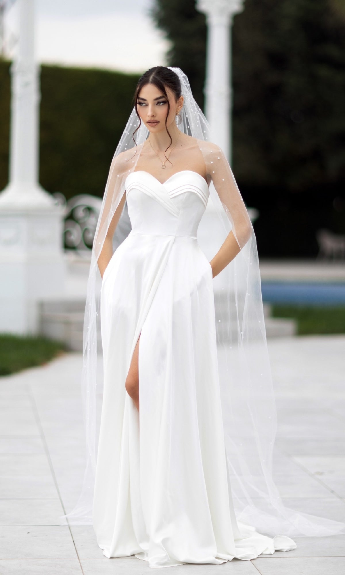 Woman in the Off-White Strapless Bridal Gown: Tina Holly TW105, featuring a thigh-high slit and sheer veil, stands outdoors on a paved area with greenery and columns in the background.